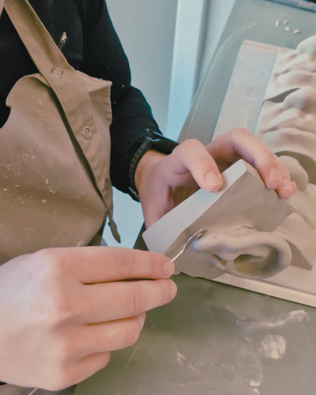 A close-up of a person's hands sculpting a detailed ear from a gray clay block, wearing a beige pottery apron over a black shirt.
