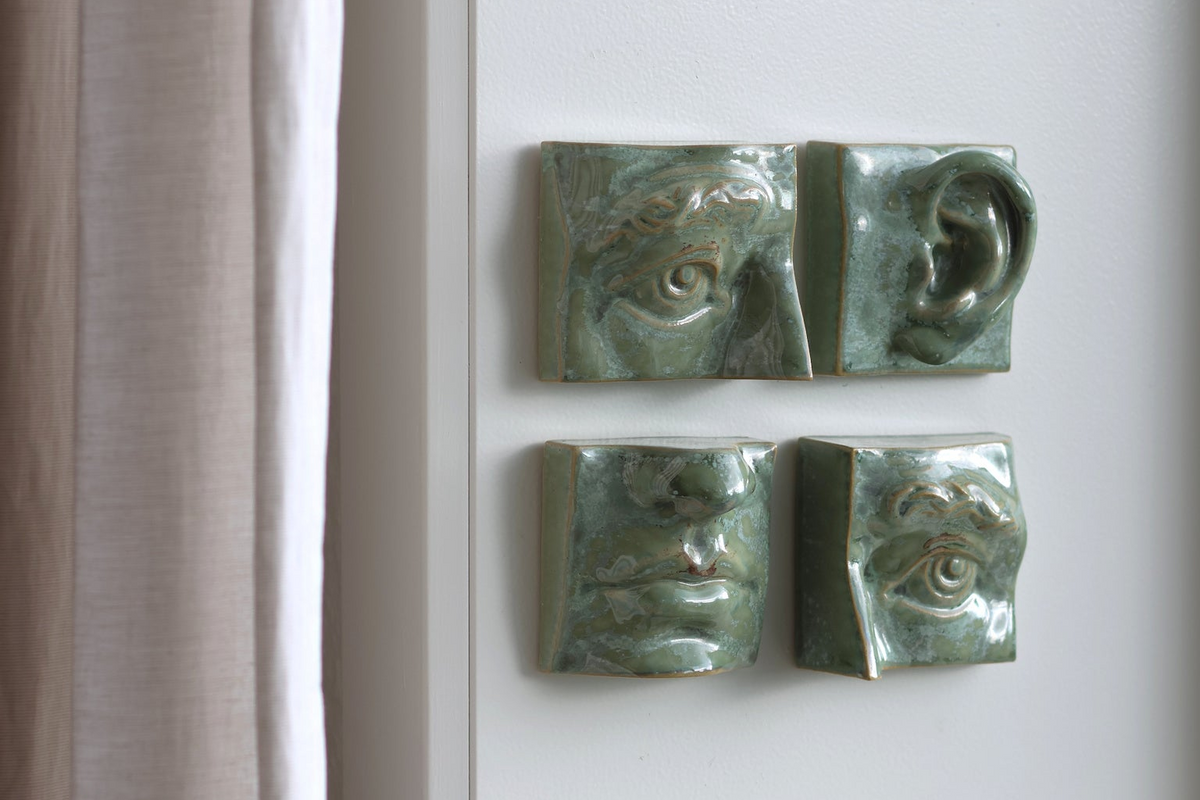 Four green glazed ceramic sculptural face fragments mounted on a white wall next to a curtain.
